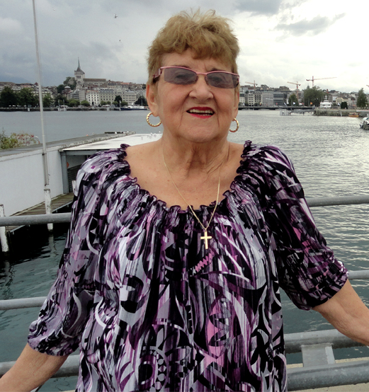 Fotografía de Ofelia Miriam Ortega, quien ha dedicado su vida a la teología feminista latinoamericana, de medio cuerpo vistiendo una blusa estampada en color violeta recostada a la baranda de un puente. Se nota agua al fondo, del otro lado una vieja ciudad.