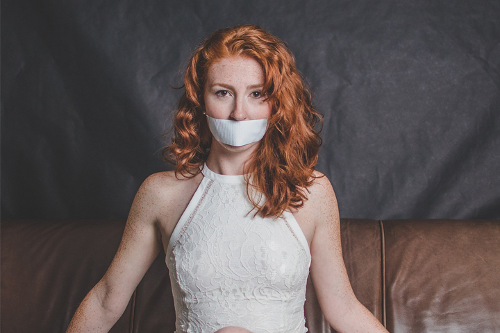 mujer pelirroja con vestido blanco sentada con la boca cubierta, significando el silencio de las mujeres